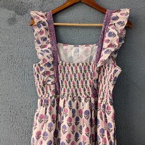 May include: A white cotton dress with a pink and blue floral print. The dress has a square neckline, ruffled straps, and a tiered skirt.