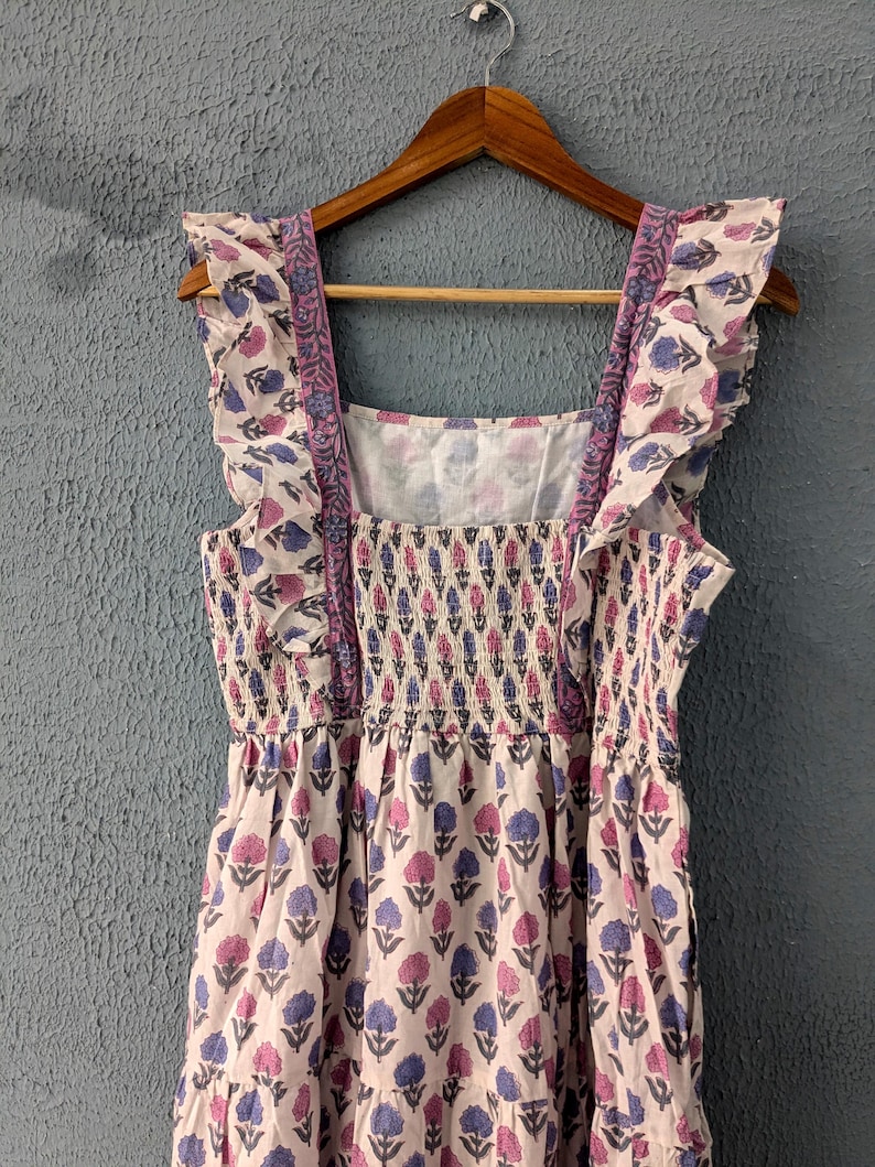 May include: A white cotton dress with a pink and blue floral print. The dress has a square neckline, ruffled straps, and a tiered skirt.