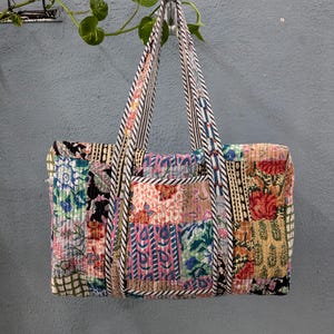 May include: A patchwork quilted tote bag with floral and geometric patterns in shades of pink, green, blue, and brown. The bag has black and white striped straps.