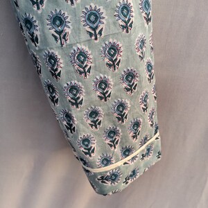 May include: A close-up of a light blue fabric with a white trim. The fabric has a repeating pattern of blue and green floral designs.
