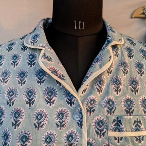 May include: A light blue pajama top with a white trim and a floral print. The top has a button closure and a pocket on the left side.