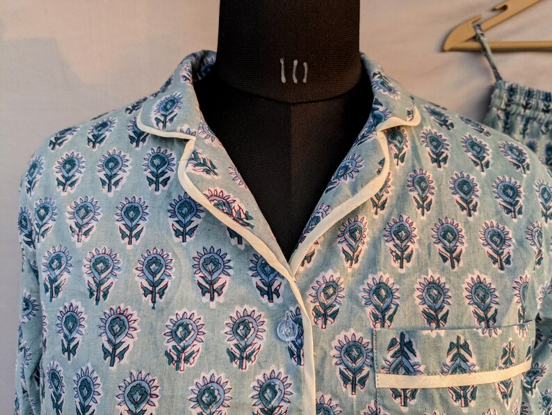 May include: A light blue pajama top with a white trim and a floral print. The top has a button closure and a pocket on the left side.