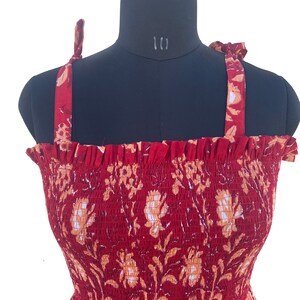 May include: A red sundress with a floral pattern, displayed on a mannequin. The dress features a smocked bodice and tie straps. The floral design includes white and orange flowers and leaves.