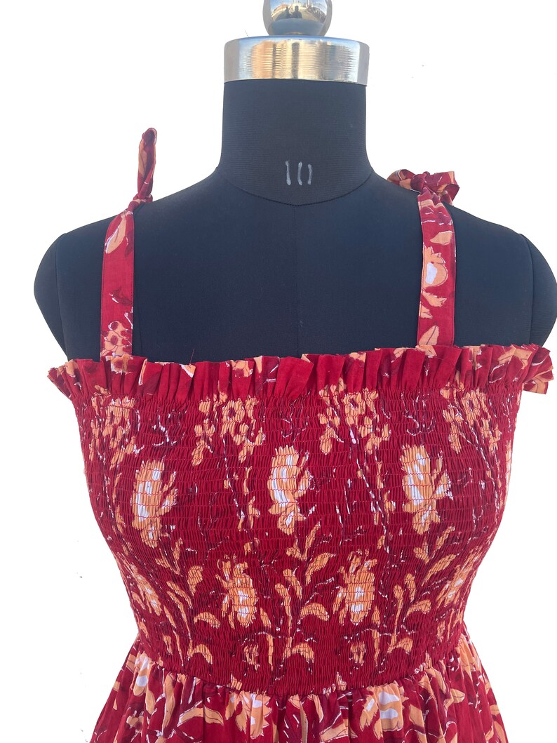 May include: A red sundress with a floral pattern, displayed on a mannequin. The dress features a smocked bodice and tie straps. The floral design includes white and orange flowers and leaves.