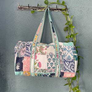 Puede incluir: Una bolsa de viaje estilo patchwork con fondo blanco y estampados florales coloridos. La bolsa tiene un ribete a rayas blanco y azul claro y dos asas.