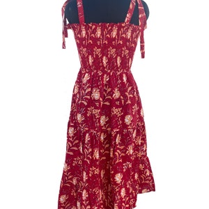May include: A red sundress with a floral pattern. The dress features tie straps, a smocked bodice, and a tiered skirt. The floral design includes white and peach-colored flowers and leaves. The dress is displayed on a black mannequin.
