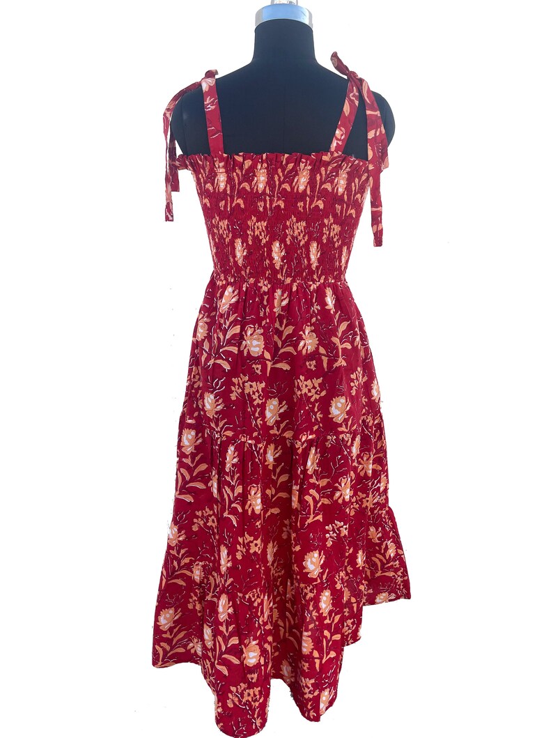 May include: A red sundress with a floral pattern. The dress features tie straps, a smocked bodice, and a tiered skirt. The floral design includes white and peach-colored flowers and leaves. The dress is displayed on a black mannequin.