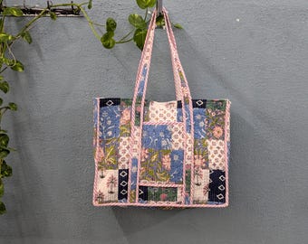 May include: A patchwork tote bag with a pink and blue floral pattern. The bag has a pink and white trim and is hanging on a hook.