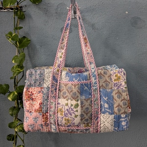 May include: A patchwork quilted duffel bag with pink, blue, and white floral patterns. The bag has a pink and white striped handle and a small pocket on the front.