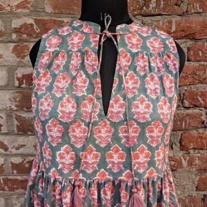 May include: A sleeveless green dress with a pink floral print and a tie at the neck. The dress has a tiered skirt and pink tassels.