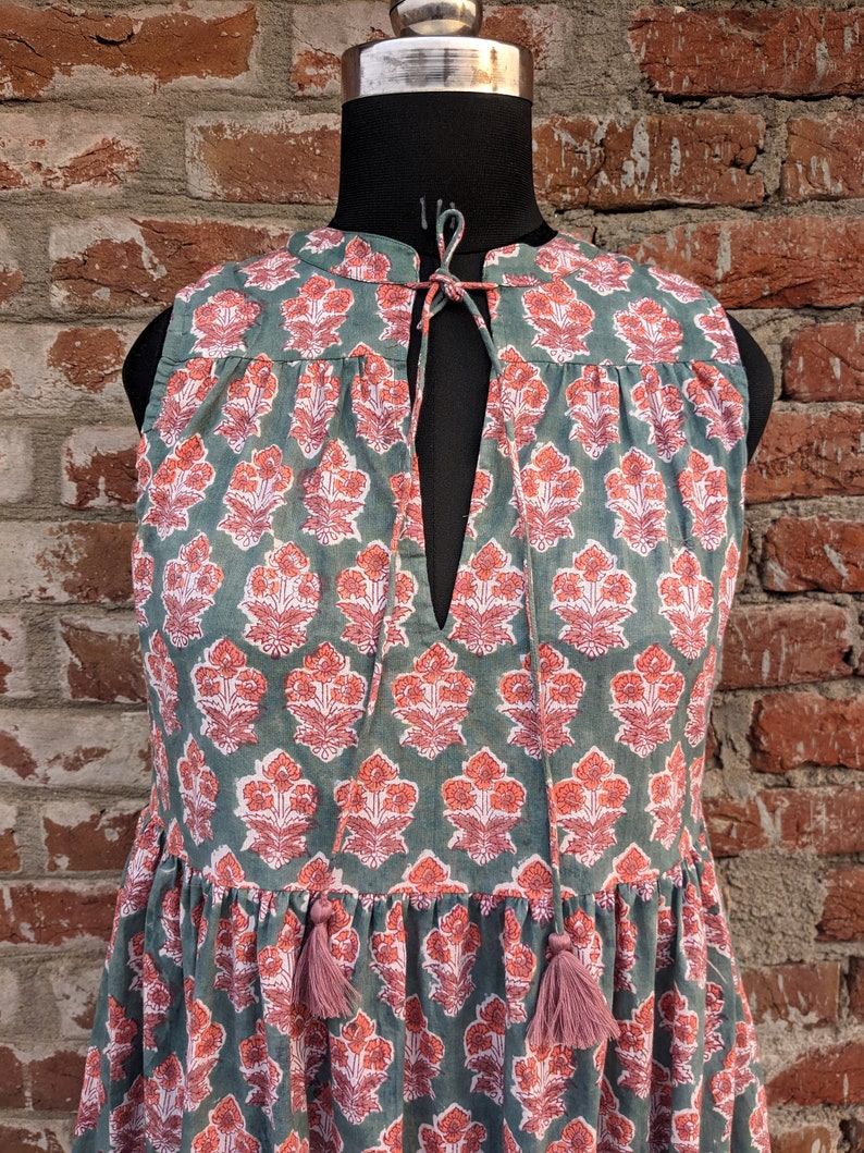 May include: A sleeveless green dress with a pink floral print and a tie at the neck. The dress has a tiered skirt and pink tassels.