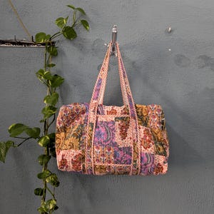 Op de afbeelding: Een roze en paarse quilted reistas met een bloemenpatroon en een zilveren haak.