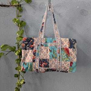 May include: A patchwork style tote bag with a blue, beige, and green floral print. The bag has white and black striped straps and is hanging on a hook.