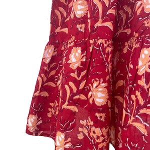 May include: A section of a red fabric with a floral pattern. The design features large, stylized flowers in shades of peach and white, set against a deep red background. The fabric has a tiered, ruffled edge.