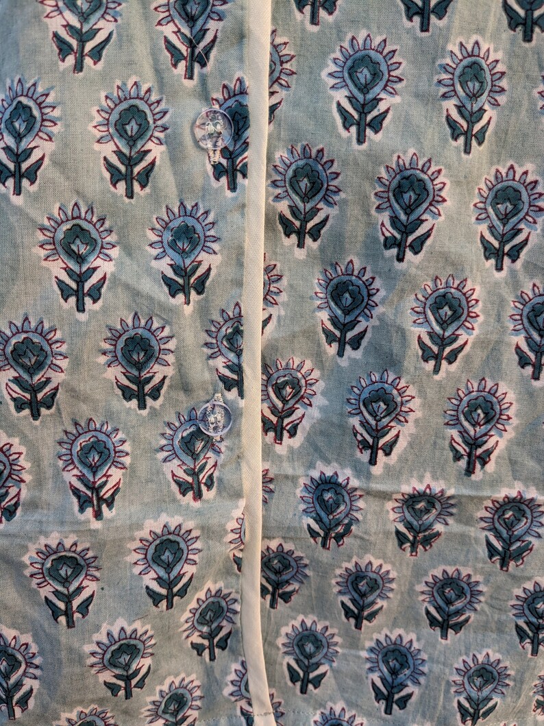May include: A close-up of a light blue fabric with a repeating pattern of small, stylized floral designs in shades of blue, white, and red. The fabric has a white seam running down the center.