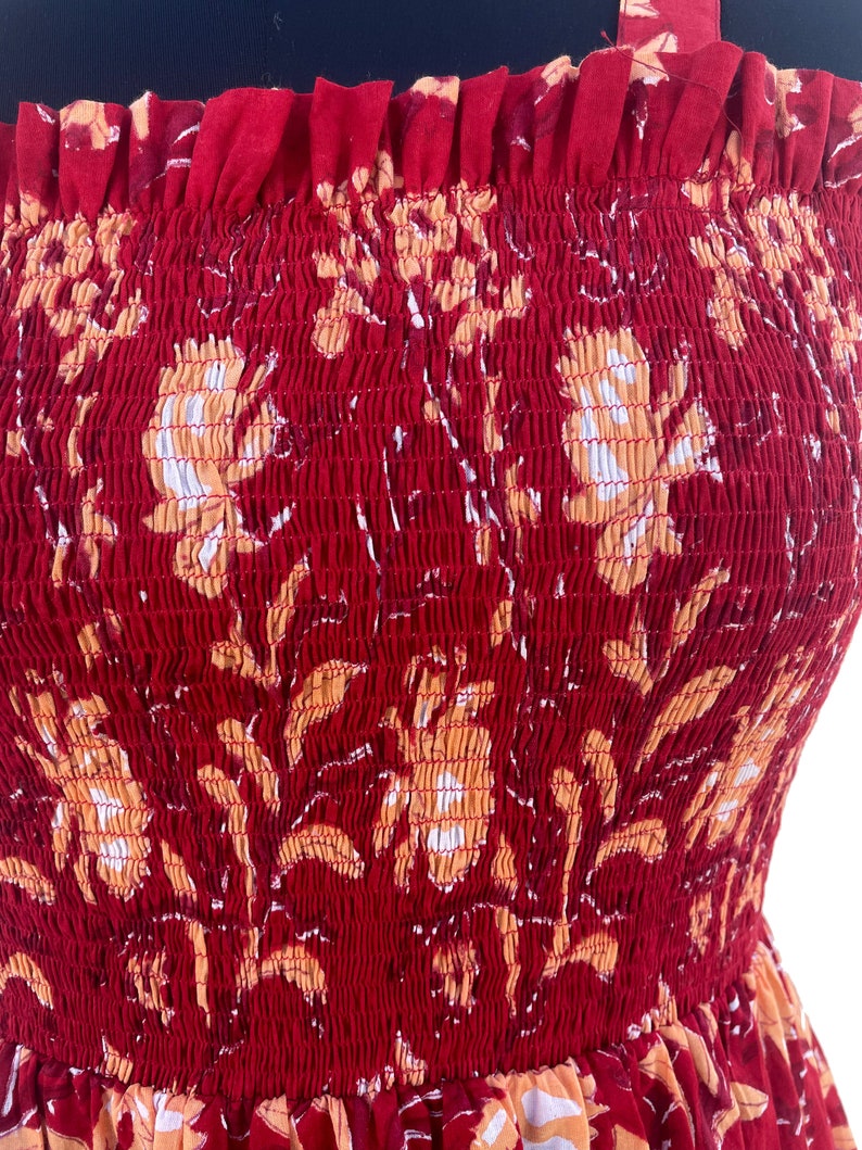 May include: A red sundress with a floral pattern in shades of orange and white. The dress features a smocked bodice and thin straps. The fabric has a textured appearance.