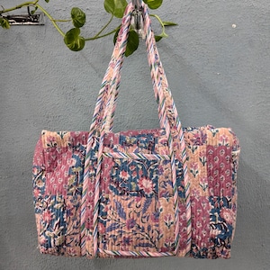 May include: A pink and blue floral patterned quilted tote bag with a long strap. The bag is hanging on a hook.