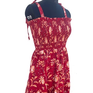 May include: A red sundress with a floral pattern, displayed on a black mannequin. The dress features tie straps and a smocked bodice. The floral design includes orange and white accents.