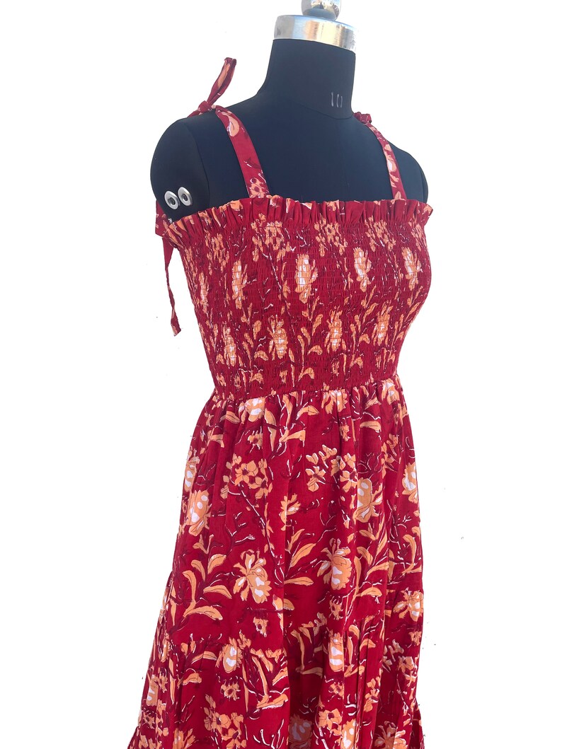 May include: A red sundress with a floral pattern, displayed on a black mannequin. The dress features tie straps and a smocked bodice. The floral design includes orange and white accents.