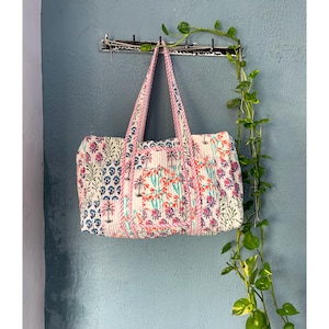 Könnte beinhalten: Eine weiße gesteppte Tragetasche mit pinkfarbenem und blauem Blumendruck. Die Tasche hat zwei Henkel und hängt an einem Haken.