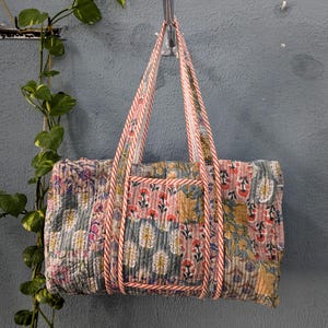 May include: A quilted floral print tote bag with red and white striped handles. The bag is hanging on a hook against a gray wall.