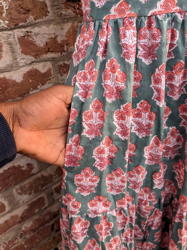 May include: A green cotton dress with a floral print in shades of pink and white. The dress has a gathered skirt and a pocket on the side.