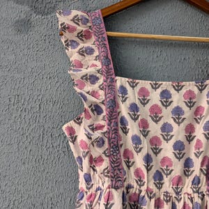 May include: A white cotton dress with a pink and blue floral print. The dress has a square neckline, ruffled straps, and a gathered waist.