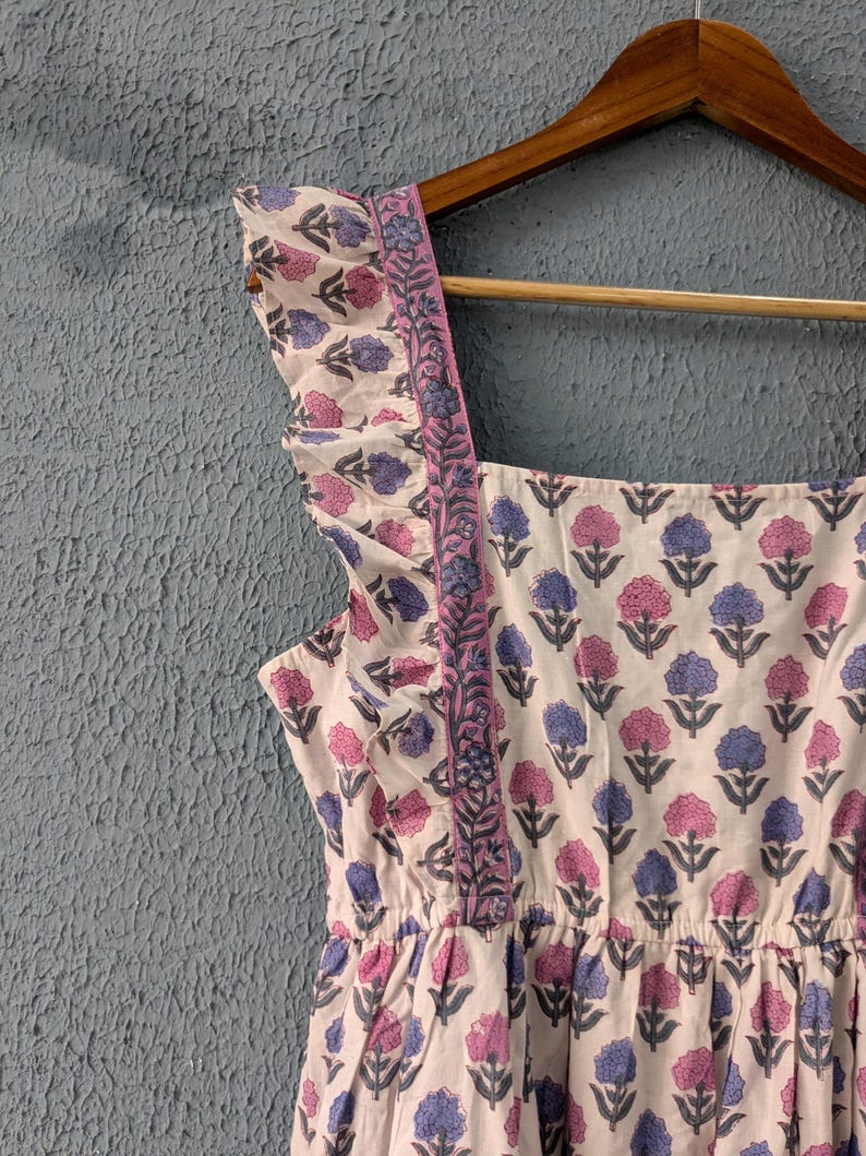 May include: A white cotton dress with a pink and blue floral print. The dress has a square neckline, ruffled straps, and a gathered waist.
