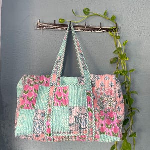 Könnte beinhalten: Eine Dufflebag im Patchwork-Stil mit rosa und grünen Blumenmustern. Die Tasche hat zwei Träger und hängt an einem Haken.