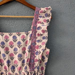 May include: A white cotton dress with a floral print in shades of pink and blue. The dress has a square neckline, ruffled straps, and a gathered skirt.