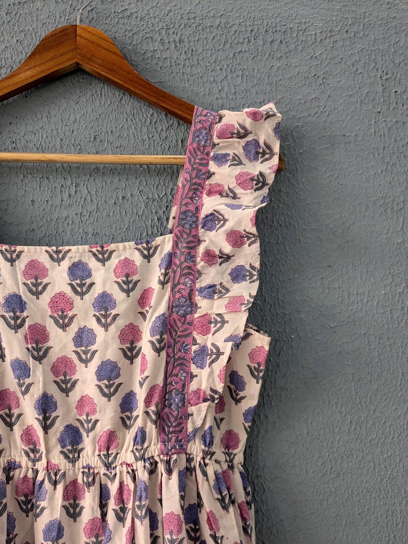 May include: A white cotton dress with a floral print in shades of pink and blue. The dress has a square neckline, ruffled straps, and a gathered skirt.