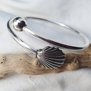 Peut inclure: Un bracelet en argent avec un charme en forme de coquille et une boule à l'extrémité. Le bracelet est posé sur un morceau de bois flotté.