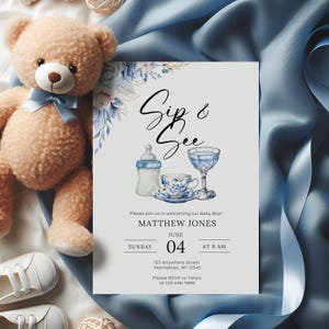 Op de afbeelding: Een uitnodiging voor een babyshower met de tekst "Sip & See" en details voor de babyjongen van Matthew Jones. De uitnodiging bevat aquarel illustraties van een babyfles, een kopje en een glas. Een pluche teddybeer en blauwe stof omringen de uitnodiging.