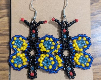 May include: A pair of butterfly-shaped dangle earrings crafted from small seed beads. The wings feature a pattern of bright yellow and translucent blue beads, while the body is constructed from black beads with small red accents at the tips.