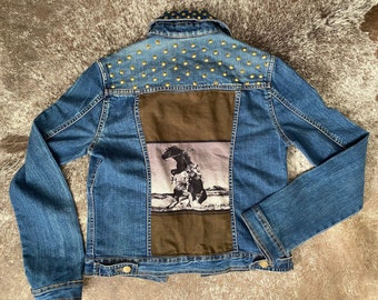 May include: A blue denim jacket with gold studs on the back. The back of the jacket has a brown patch with a black and white image of a person riding a horse.