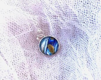 May include: A round pendant necklace with a silver chain. The pendant has a blue, gold, and white abstract design set in a silver bezel. The necklace is displayed on a white, textured fabric.