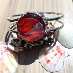 May include: A silver-toned bracelet with a large, round, red and white abstract design. The bracelet is displayed on a seashell, with other seashells in the background. The design has a marbled effect.