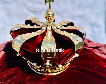 May include: A gold crown with a red velvet lining and white pearl accents. The crown has a cross on top and is decorated with green gemstones.