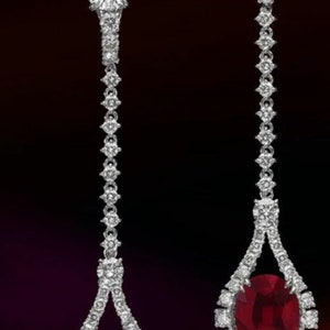 May include: A pair of long, dangling earrings with a teardrop-shaped design. Each earring features a large, oval-shaped ruby surrounded by a halo of smaller diamonds. The earrings are made of silver and have a delicate, elegant look.