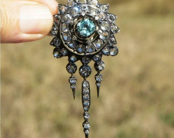 May include: A silver and diamond brooch with a large blue gemstone in the center. The brooch has a circular design with a dangling chain of diamonds.
