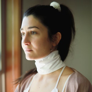 May include: A woman with dark hair tied back in a ponytail with a white scrunchie, wearing a white turtleneck and a light brown cardigan. She is looking off to the side.
