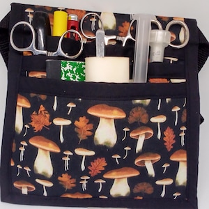 Peut inclure: Une ceinture utilitaire en tissu noir avec plusieurs poches, ornée d'un motif de champignons et de feuilles. Les poches contiennent divers outils, dont des ciseaux, une seringue et d'autres fournitures médicales. La ceinture a une sangle noire réglable.