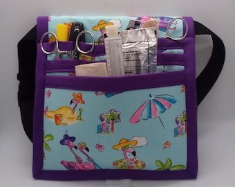 May include: A purple and blue medical utility belt with multiple pockets. The belt has a flamingo and palm tree print. The belt is filled with medical supplies, including scissors, a ruler, and a tube of ointment.