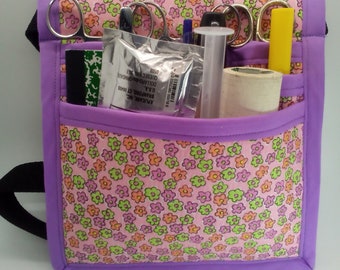 May include: Purple medical utility bag with a pink floral print. The bag is filled with medical supplies, including scissors, a tube, a roll of tape, and a yellow highlighter.