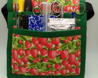 May include: A green fabric tool belt with a strawberry print. The belt has multiple pockets and loops for holding tools and supplies. The belt is filled with various tools, including scissors, tweezers, a yellow highlighter, a blue pen, and a clear plastic tube.