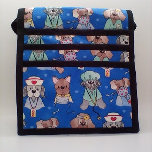 Pu&ograve; includere: Una borsa in tessuto blu con un bordo nero e un motivo ripetuto di cani dei cartoni animati che indossano camici medici e maschere. I cani indossano cappelli, tengono stetoscopi e alcuni hanno bende. La borsa ha una tracolla nera e le parole "LOVE" e "VET" sono visibili sul tessuto.
