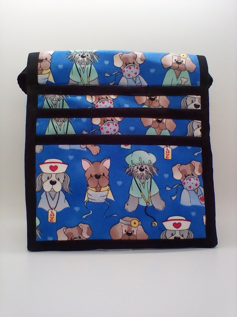 Pu&ograve; includere: Una borsa in tessuto blu con un bordo nero e un motivo ripetuto di cani dei cartoni animati che indossano camici medici e maschere. I cani indossano cappelli, tengono stetoscopi e alcuni hanno bende. La borsa ha una tracolla nera e le parole "LOVE" e "VET" sono visibili sul tessuto.