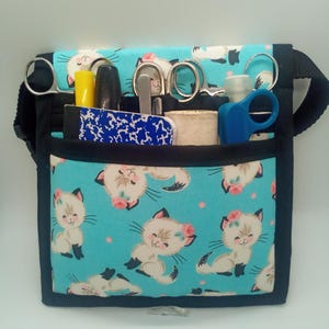 May include: A turquoise fabric utility pouch featuring a repeating pattern of cartoon kittens with pink flowers. The pouch has black trim, an adjustable strap, and multiple exterior pockets containing various tools like scissors and markers.