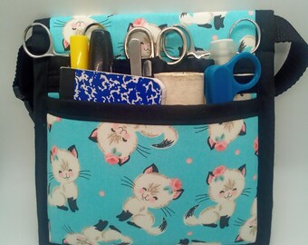May include: A turquoise fabric utility pouch featuring a repeating pattern of cartoon kittens with pink flowers. The pouch has black trim, an adjustable strap, and multiple exterior pockets containing various tools like scissors and markers.