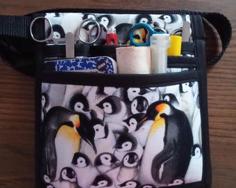 Può includere: Un marsupio con bordi neri caratterizzato da un motivo ripetuto di pinguini adulti e pulcini. La borsa è dotata di diverse tasche frontali contenenti vari strumenti, tra cui forbici in metallo, un piccolo taccuino e contenitori cilindrici.